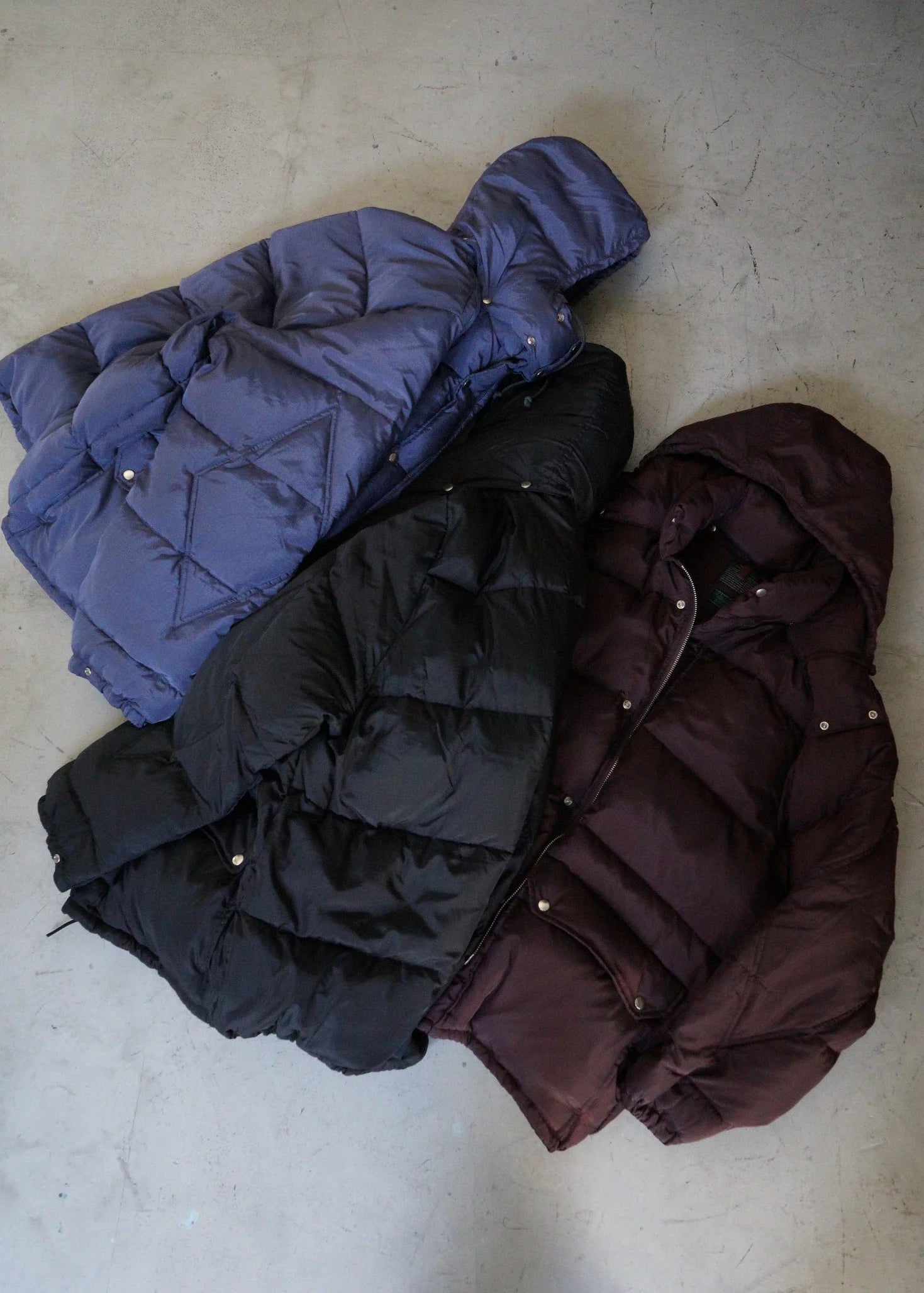 Three puffer jackets in purple, black, and maroon on a light gray surface.