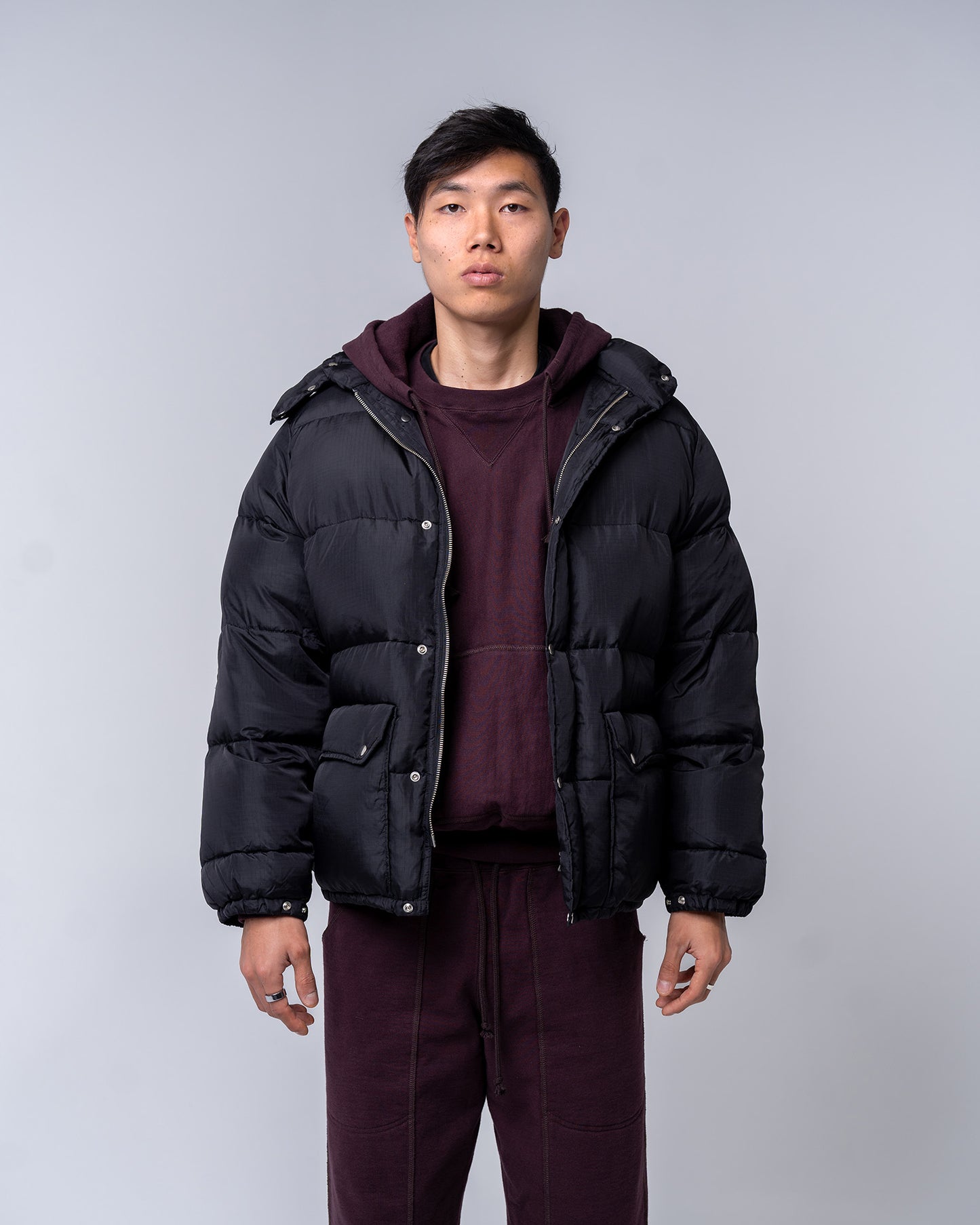 Person wearing a black puffer jacket over a maroon hoodie and pants on a gray background