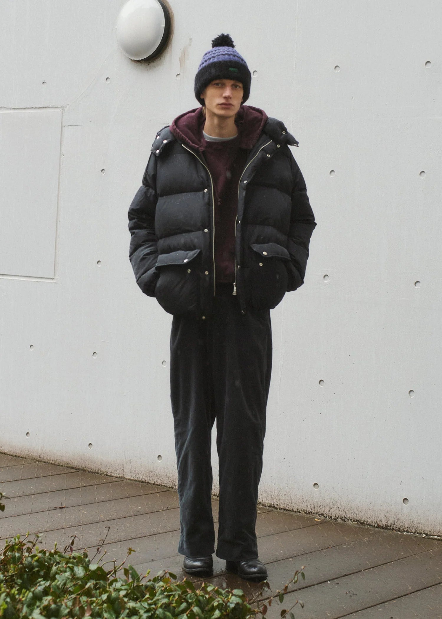 Person wearing a black puffer jacket and dark pants standing against a white wall.