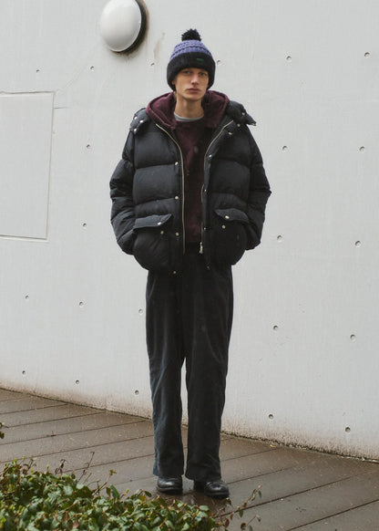 Person wearing a black puffer jacket and dark pants standing against a white wall.