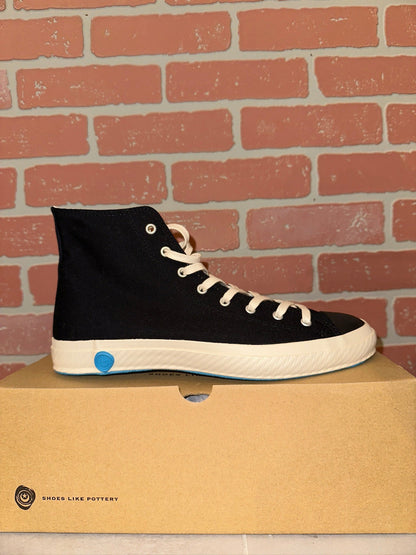 Black high-top sneaker with white laces on a cardboard box against a brick wall background